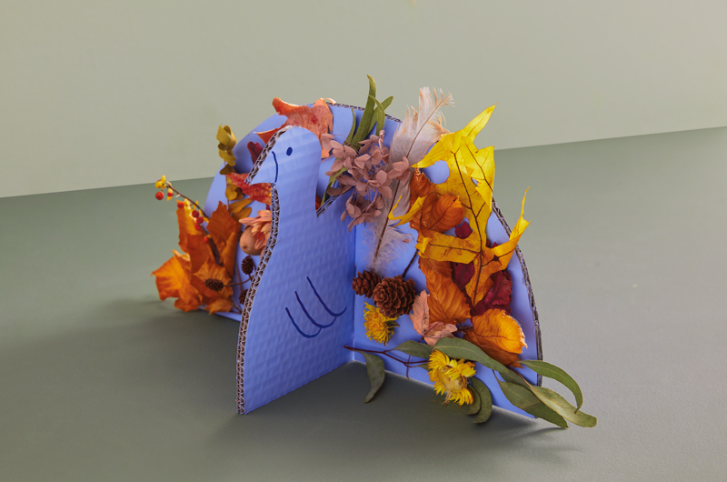 A Thanksgiving centerpiece shaped like a turkey; the body and tail of the turkey are made out of cardboard and painted blue; the tail is decorated with foraged fall items like fallen leaves, dried flowers and acorns.