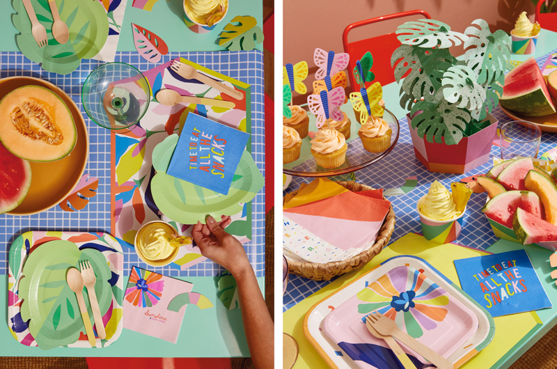 A table is set for a tropical theme party using various partyware products and table decorations, including a paper centerpiece made to look like a monstera plant in a pot, paper butterfly cupcake toppers, leaf-shaped paper plates, and brightly colored snack trays, cups and napkins.
