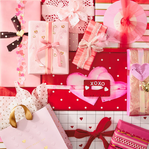A collection of gifts wrapped with paper in shades of red, pink, gold and white, some with contrasting stripes, others with hearts and polka dots; many gifts are tied with bows made of ribbon.