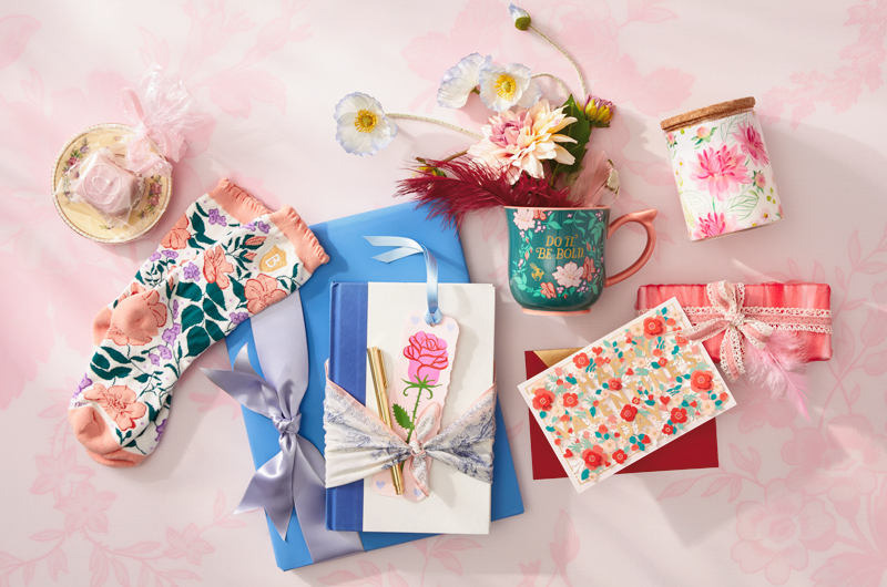 A collection of gift ideas for a book lover on Galentine's Day, including a journal with pen, a Bridgerton tea mug and pair of cozy socks, a floral tea canister, a floral trinket dish, and a papercraft card that reads