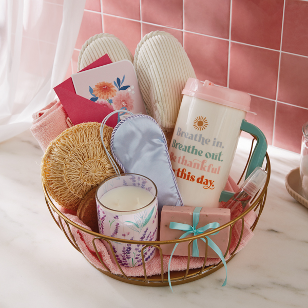 A spa-themed Mother's Day gift basket that includes an eye mask, scented candle, a loofah, a pair of slippers, handmade soap, a floral card, and in insulated tumbler that reads, 