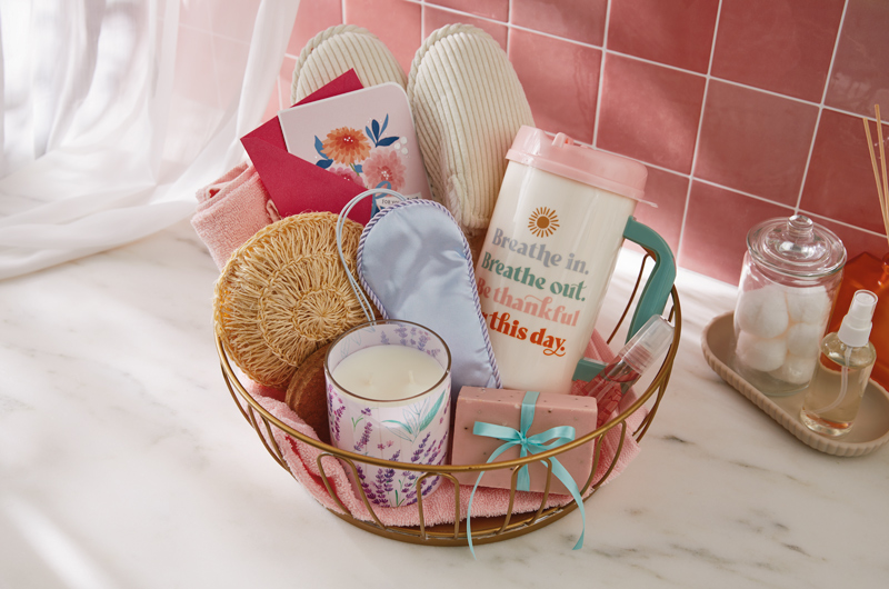A spa-themed Mother's Day gift basket that includes an eye mask, scented candle, a loofah, a pair of slippers, handmade soap, a floral card, and in insulated tumbler that reads, 