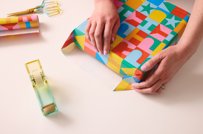 A woman's hands fold down the edges of wrapping paper along the side of a present being wrapped.