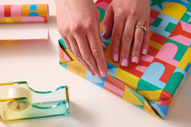 A woman's hands fold up the edge flap of a gift that she is wrapping; a roll of tape sits ready nearby.