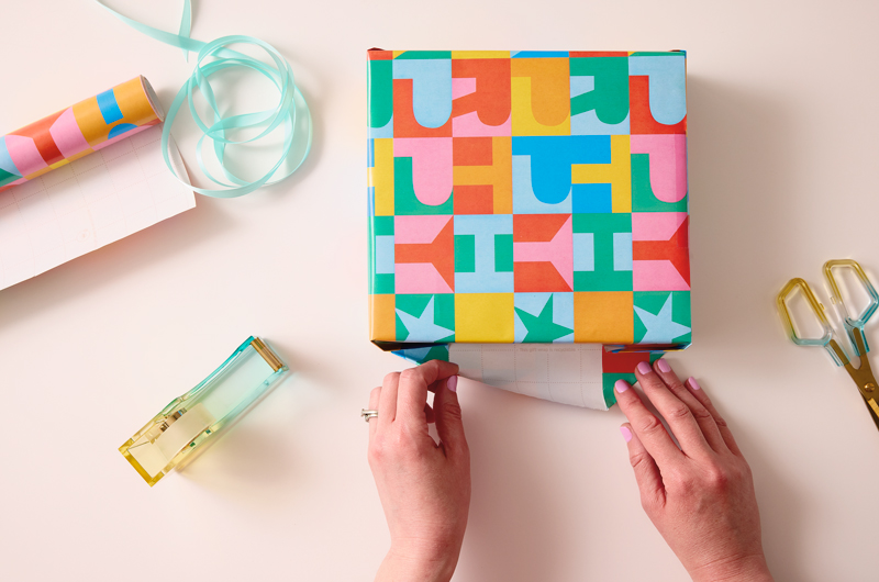 A woman's hands are shown folding up a flap on the edge of a gift that she is wrapping; a roll of tape sits ready nearby.