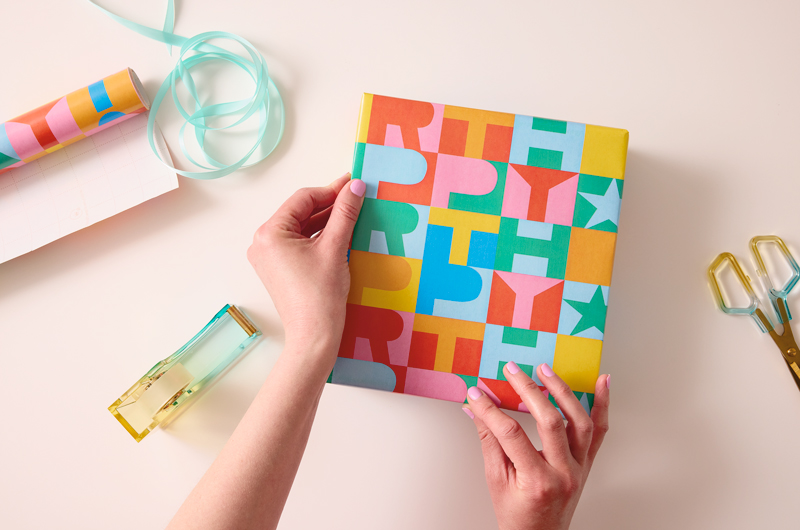 A woman uses her forefinger and thumb to gently crease wrapping paper along the top corners of a gift she has wrapped, to make the gift presentation a little neater.