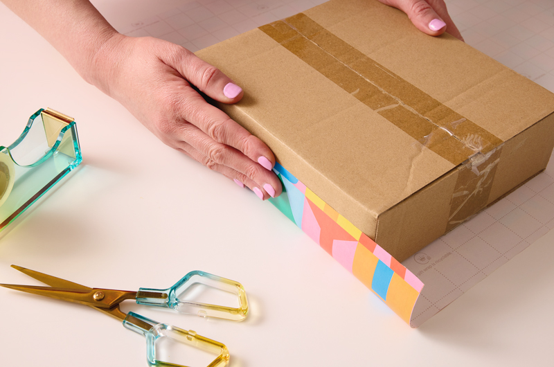 A woman's hands are shown placing a box on top of a piece of wrapping paper for the purpose of measuring out how much wrapping paper she needs to wrap the box.