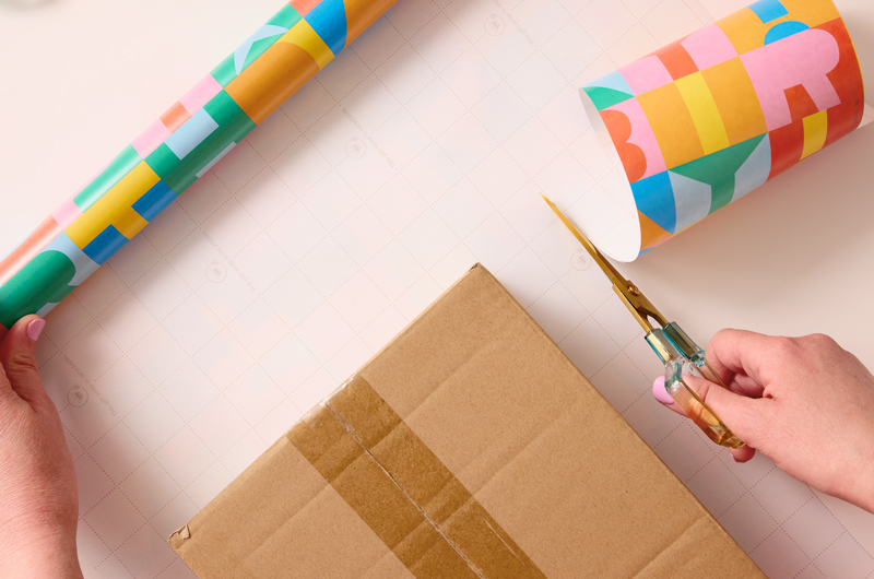 A woman's hands cut wrapping paper alongside a cardboard box that's intended to be wrapped.