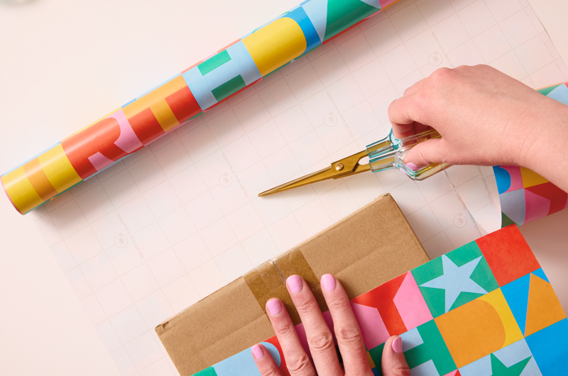 A woman's hand holds a pair of scissors and indicates where on the reverse of the gift wrap--which is printed with grid lines to assist in measuring and cutting--where she intends to cut the wrap, width-wise.