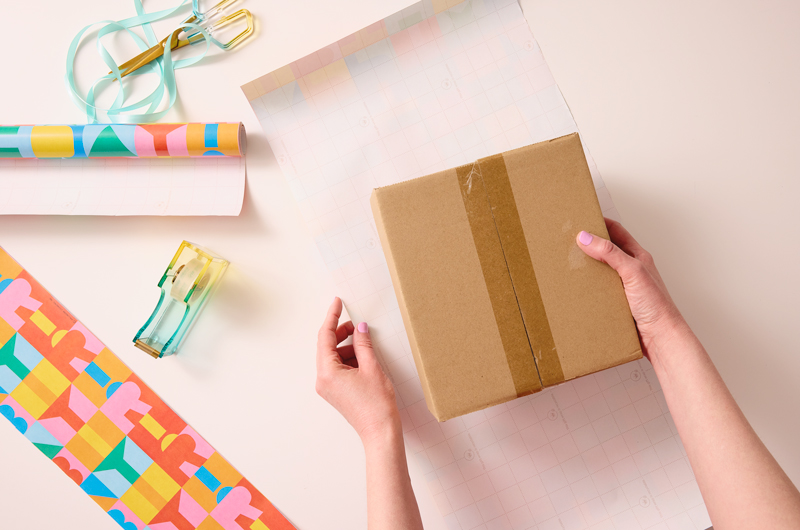 A woman's hands place a plain cardboard box on top of a piece of gift wrap that she has measured to fit her needs.