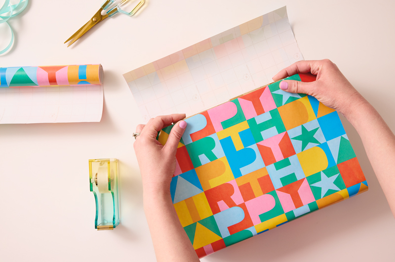 A woman's hands fold one end of a strip of wrapping paper over a box.