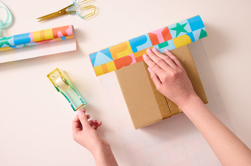 A woman holds down a piece of wrapping paper on top of a cardboard box while reaching for a piece of tape with her other hand.