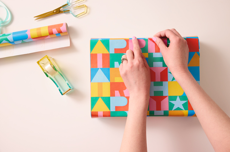 A woman's hands smooth down a piece of tape that she has used to secure two ends of a piece of wrapping paper together on a gift box.