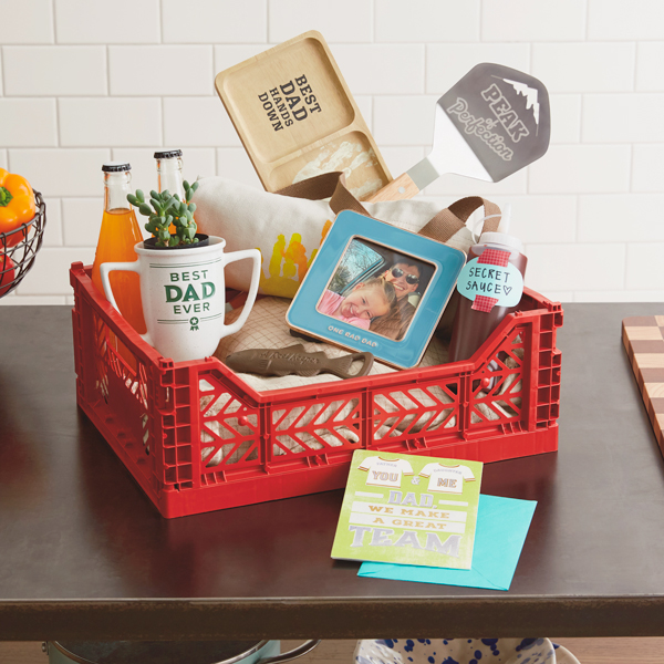 A DIY Father's Day gift basket that includes a card, a framed photo of a father and his toddler daughter, a metal bottle opener in the shape of a trout, a pencil cup in the shape of a trophy that reads,
