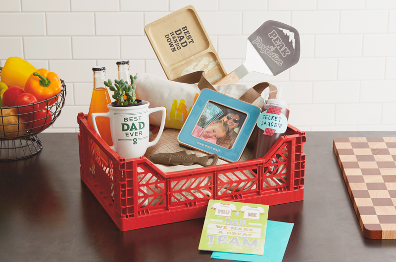 A DIY Father's Day gift basket that includes a card, a framed photo of a father and his toddler daughter, a metal bottle opener in the shape of a trout, a pencil cup in the shape of a trophy that reads,