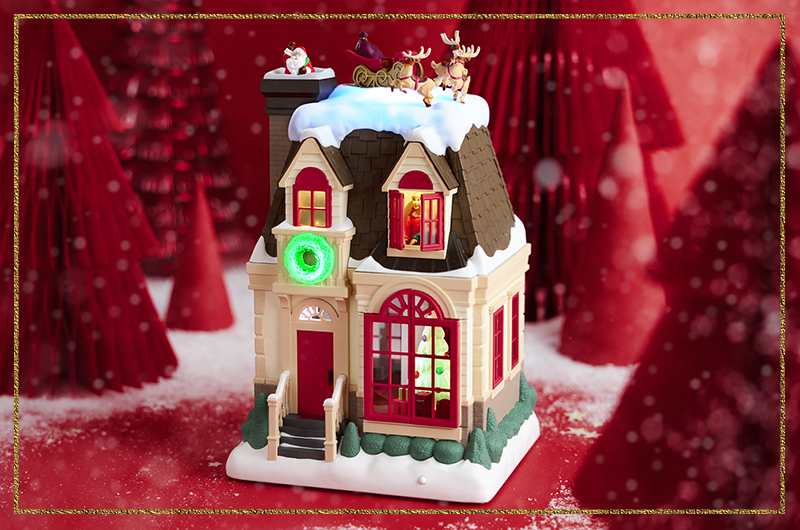 A table top figurine with light, sound and motion; the figurine is a model house that depicts several scenes, from Santa slipping down a chimney, to children waking up in their bedrooms, to the children spying Santa as he goes to put presents under a lit Christmas tree.