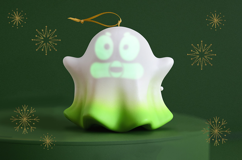 A Keepsake Ornament shaped like a ghost sits on a dark green platform against a dark green background; the ghost is lit up from within, revealing a green, glowing face.