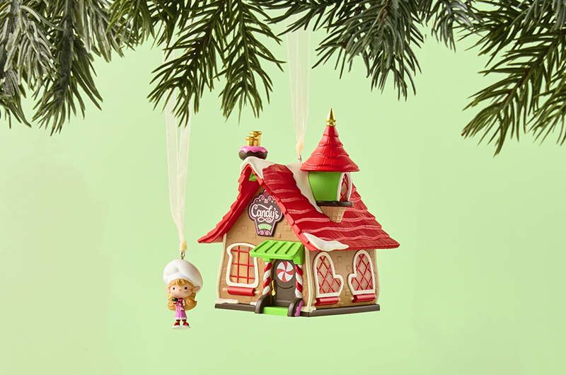 A set of two Keepsake Ornaments hangs from a Christmas tree branch; one ornament is a little elf in a chef's hat and apron, holding out a cupcake; the other