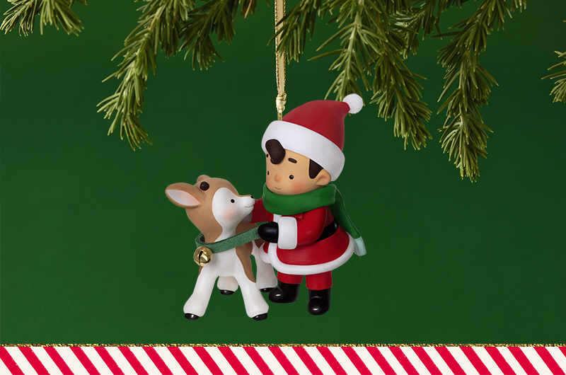A Keepsake Ornament in the Young Santa series; a little boy in a Santa outfit, understood to be Santa as a child, puts a jingle bell collar on a baby reindeer.