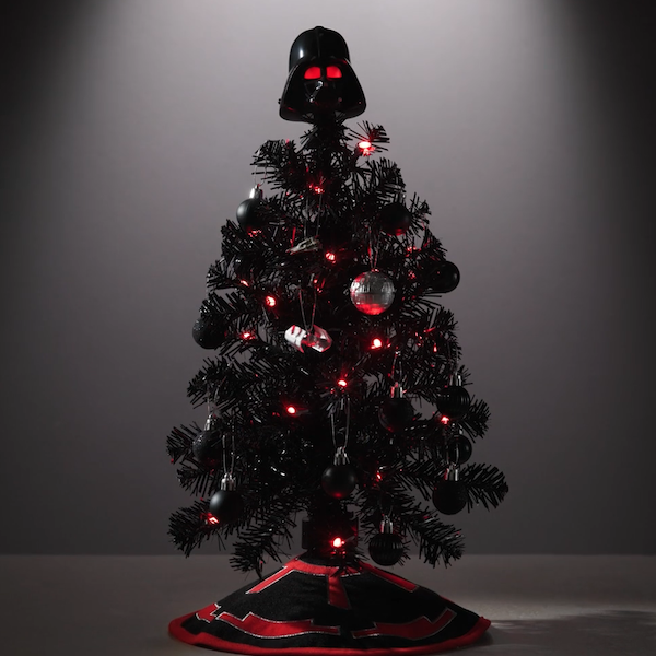 A black miniature Christmas tree is topped with a Star Wars Darth Vader helmet that lights up from within and coordinates in color with the lights on the tree; the lights glow red and there is a red fabric tree skirt at the base of the mini tree.
