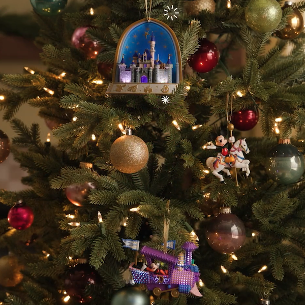 A trio of Disney Parks-themed Keepsake Ornaments hangs on a decorated Christmas tree; the ornaments feature a scene of Sleeping Beauty's Castle against a night sky, Goofy's train from the Main Street Electrical Parade, and Mickey riding a horse from King Arthur's Carrousel.