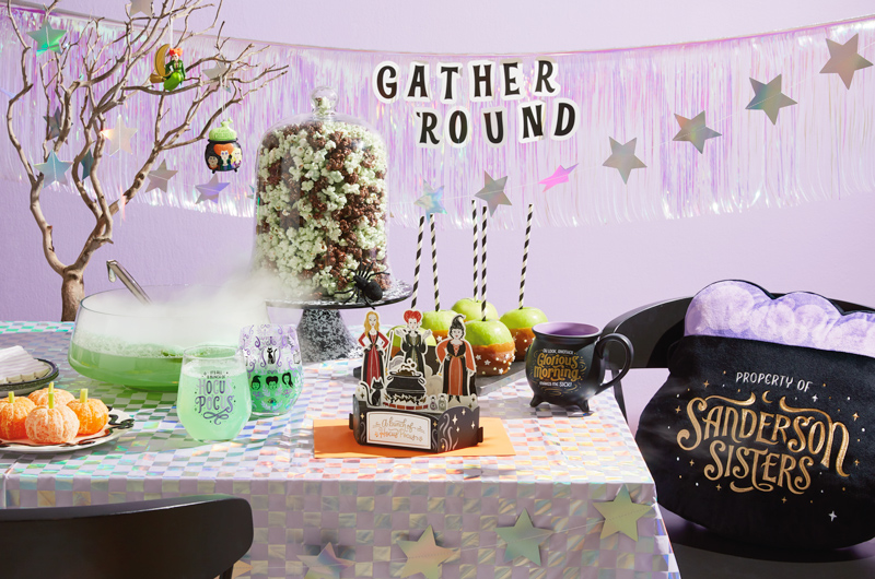 A party table set up for a Disney Hocus Pocus-themed Halloween party; the table includes a light-up pillow shaped like a bubbling cauldron that reads,