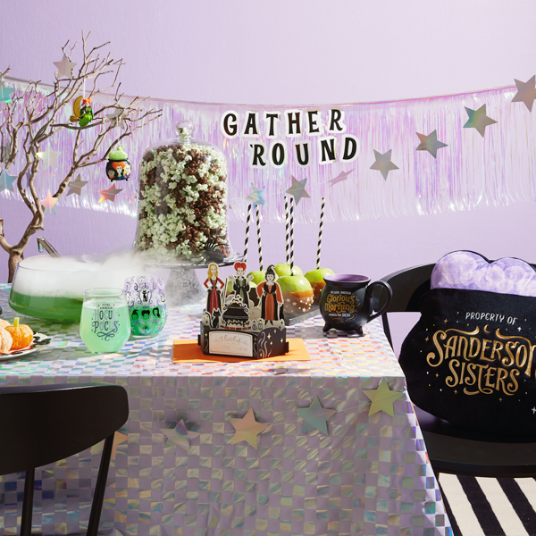 A party table set up for a Disney Hocus Pocus-themed Halloween party; the table includes a light-up pillow shaped like a bubbling cauldron that reads,