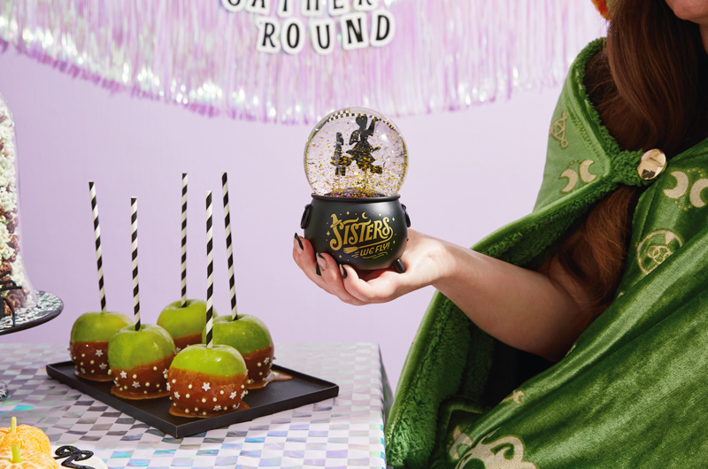 A woman holds a Disney Hocus Pocus snow globe that features a silhouette of the Sanderson Sisters inside, along with gold glitter; beneath the glass globe is a black cauldron that says,