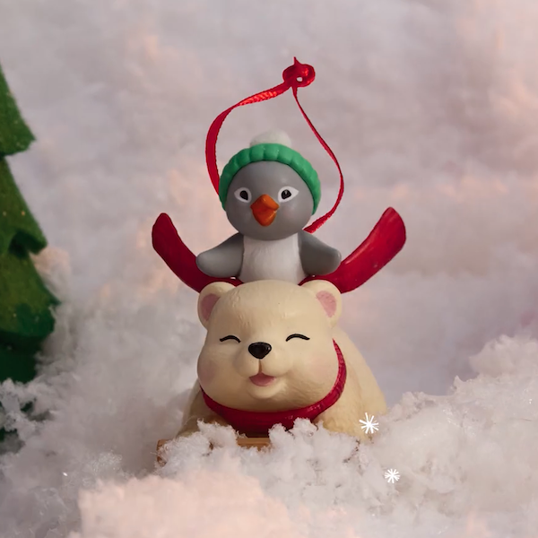 A Keepsake Ornament featuring a penguin riding on the back of a polar bear as he sleds on his belly down a snowy hillside.