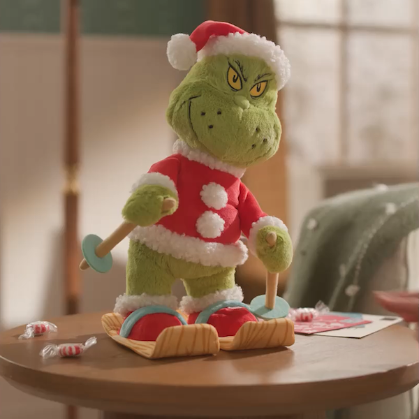 A plush version of Dr. Seuss's The Grinch is dressed in a Santa costume and stands on a pair of skis while holding two ski poles; the plush is standing on a light wood side table scattered with peppermint candies.