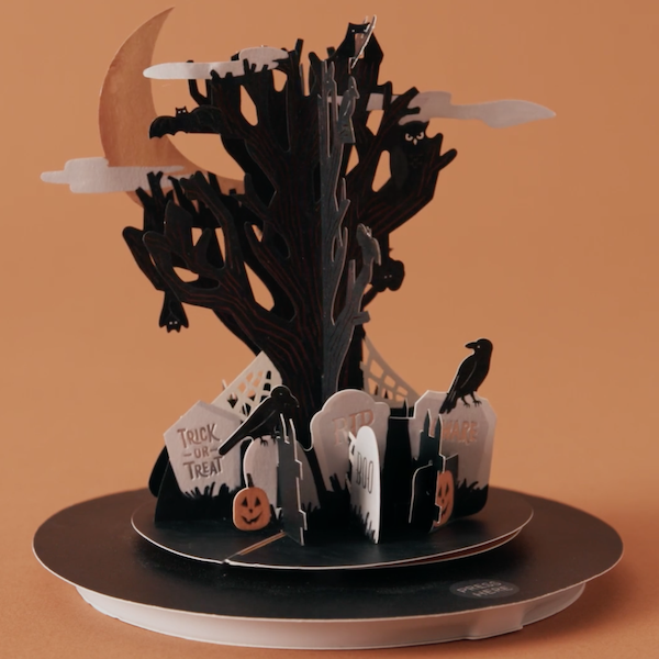 A musical, 3D pop-up card portraying a creepy, moonlit graveyard scene sits in front of an orange background.