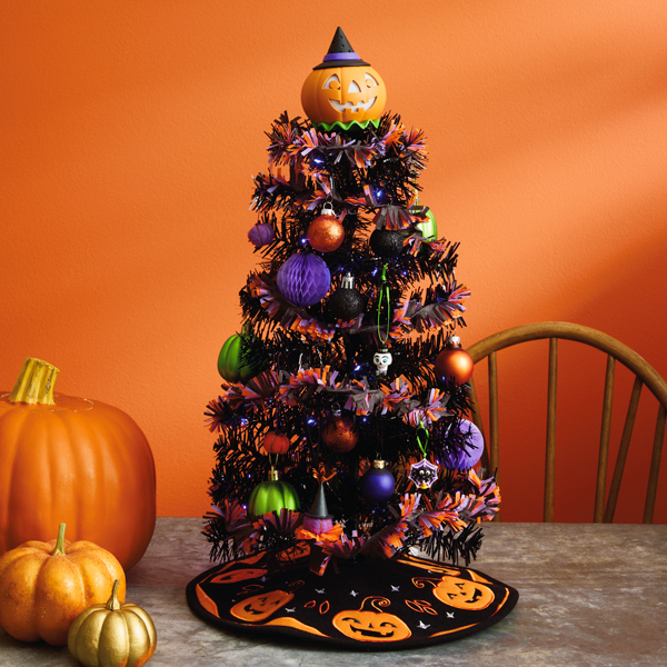 A miniature Halloween tree sits on a gray tabletop next to a trio of different sized orange pumpkins; the Halloween tree is black and is hung with mini ornaments and small glass ball ornaments in shades of purple, green and orange; the tree is topped with a jack-o'-lantern tree topper and has a little black tree skirt embroidered with orange felt jack-o'-lanterns at its base.