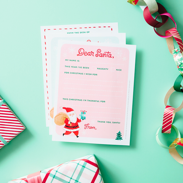 Our free, printable templates for writing a letter to Santa lay fanned out on a mint green surface, surrounded by wrapped Christmas presents and a paper chain made out of wrapping paper.