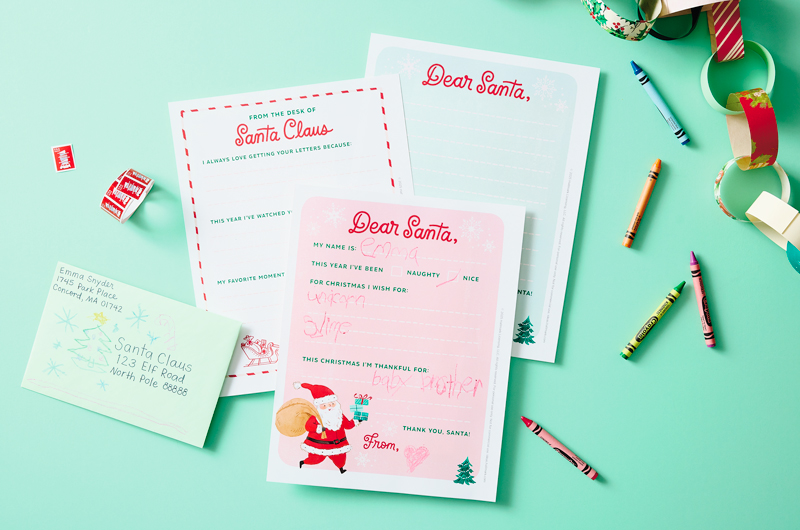 Our free, printable templates for writing a letter to Santa lay fanned out over a mint green surface; one of the templates features the childish scrawl of a little girl named Emma; scattered around the printed templates are a handful of crayons in different colors, a role of holiday postage stamps and an envelope addressed to Santa and decorated with a child's drawing of a Christmas tree.