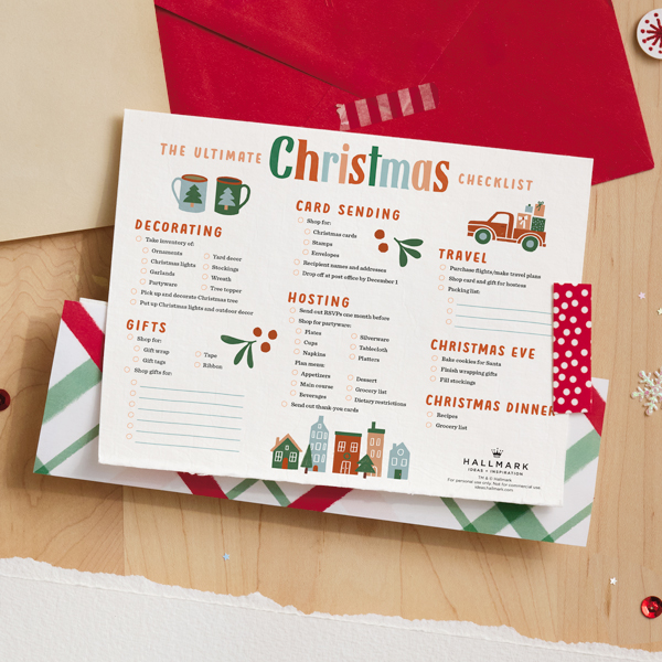 Our free printable Christmas checklist sits on a light wooden desktop with a red envelope, some wrapping paper, and bits of snowflake confetti and red spangles.