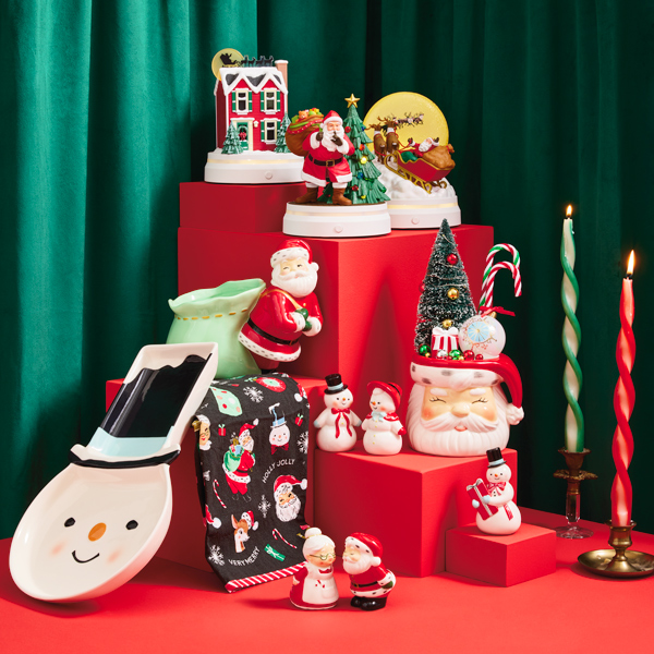 A collection of vintage-inspired decor and gifts sits on a red table top with a stair-step riser against a deep green velvet curtain background; the decor and gift items include a serving platter shaped like a snowman wearing a stove-top hat; a 3-piece set of figurines with light and sound that depict the events of the classic Christmas poem,
