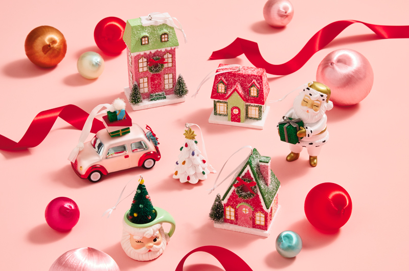 A collection of vintage-inspired Christmas ornaments sits on a pink surface, surrounded by pieces of red satin ribbon and satin ball ornaments.
