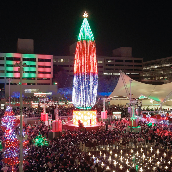 An aerial view of Crown Square during the 2024 Hallmark Christmas Experience; the square is lit up with Christmas lights and decorations; at the center of the square is an enormous Christmas tree that is several stories tall and lit up from top to bottom with lights.